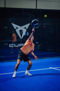 Riga, Letonya - 3 Mart 2024 - Bir oyuncu, arka planda farklı bir logo bulunan bir Padel maçında top servis ediyor.