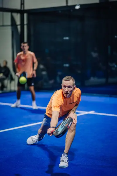 Riga, Letonya - 3 Mart 2024 - Turuncu tişörtlü odaklanmış bir oyuncu, arka planda partneri ile bir padel maçında düşük atışa hazırlanır..