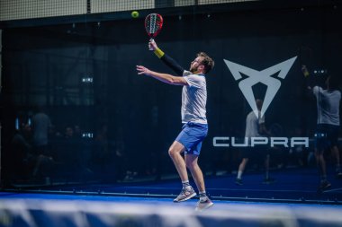 Riga, Letonya - 3 Mart 2024 - Padel maçında bir oyuncu topa vurdu, arkasında açık bir logo vardı.