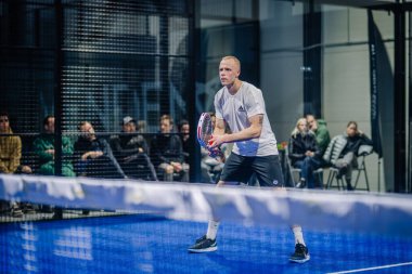 Riga, Letonya - 3 Mart 2024 - Bir adam arka planda bir izleyici ve ortağı ile vurmaya hazırlanan bir padel topuna odaklanır..