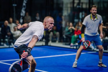 Riga, Letonya - 3 Mart 2024 - İki adam Padel oynuyor; biri topa vurmaya hazır diğeri izliyor. Seyirci arka planda görünür..
