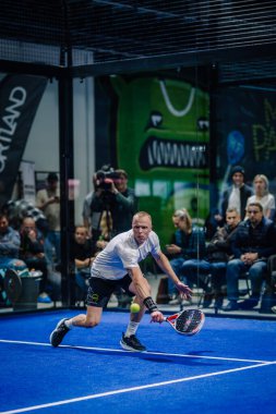 Riga, Letonya - 3 Mart 2024 - Odaklanmış bir padel oyuncusu, arka planda seyircilerin olduğu bir maç sırasında çömelmiş bir pozisyonda düşük bir topa uzanıyor.