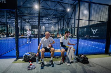 Riga, Letonya - 3 Mart 2024 - Padel Adliyesi 'ndeki bir bankta iki kişi dinleniyor, biri telefonunu kontrol ediyor, oyun devam ediyor ve arka planda seyirciler var.