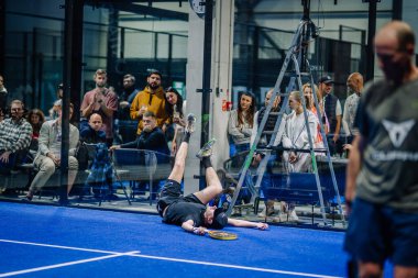Riga, Letonya - 3 Mart 2024 - Bir Padel oyuncusu sahaya düştü, ayaklar yukarıda, seyirciler camların arkasından izliyor.