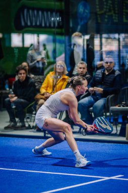 Riga, Letonya - 3 Mart 2024 - Bir padel oyuncusu beklenti içinde, raket hazır, arka planda mavi kort seyircileri ile çömelmiş.
