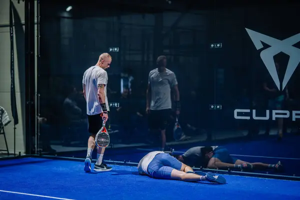 stock image Riga, Latvia - March 3, 2024 - A padel player looks on as his partner lies on the ground, possibly injured, on an indoor blue court.