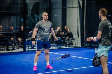 Riga, Letonya - 3 Mart 2024 - İki erkek Padel oyuncusu mavi bir kortta duruyor, biri elinde raketle, diğeri uzağa bakıyor