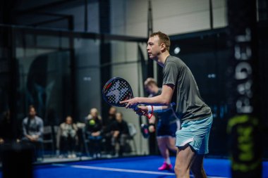 Riga, Letonya - 3 Mart 2024 - İki Padel oyuncusu oyuna odaklanıyor, biri ön planda raket tutuyor, arka planda seyirciler var.