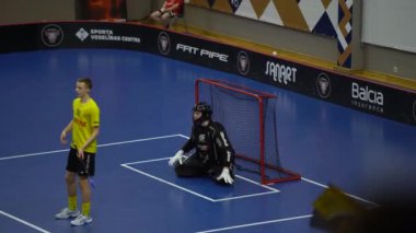 Valmiera, Letonya - 10 Mart 2024 - Kaleci siyah giyinmiş ve kale alanında diz çökmüş. Diğer oyuncular oynuyor ve hakem maçı izliyor. florbol oyunu