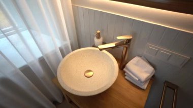 Bir banyo lavabosu, altın bir musluk ve ahşap bir vantilatör üzerinde benekli bir leğen, yanında da bir sabun makinesi, üzerinde hafif bir şerit ve havlular..