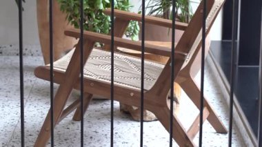 Bir ahşap direktör sandalyesi, örülmüş koltuğu ve sırtüstü, siyah metal parmaklıklardan bakıldığında arka planda bir terasotta tenceresi ve bitkisi var..
