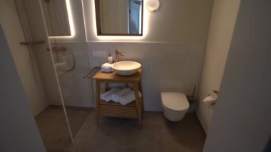 Ahşap kibrli modern bir banyo, bir tekne lavabosu, arka ışıklandırmalı bir ayna, bir duş ve duvara monte edilmiş bir tuvalet..