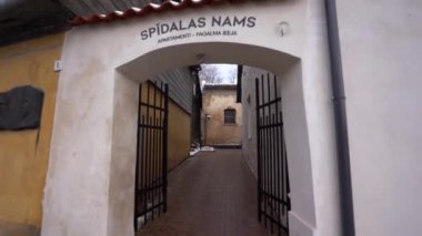 Cesis, Letonya - 14 Şubat 2024 - Sarı duvarları ve beyaz çerçeveli pencereleri olan binalarla çevrili, avluya açılan açık siyah metal kapıları olan kemerli bir giriş.