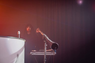 Kapüşonlu figürlü bir DJ kabini, ön planda mikrofon standı, ortam aydınlatması, performans, olay, müzik, sahne.