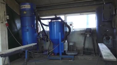 Valmiera, Letonya - 17 Mart 2024 - İyi aydınlatılmış bir odada büyük mavi bir patlama makinesi, bir kompresör, bir çalışma masası ve bir çalışma masası içeren endüstriyel ekipmanlarla dolu bir atölye.