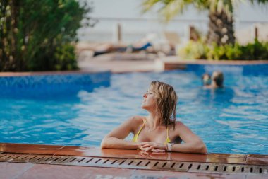 Dubai, Birleşik Arap Emirlikleri - 19 Ekim 2019 - Sarı bikinili gülümseyen bir kadın havuzun kenarına yaslanıyor, arka planda güneşlik ve palmiye ağaçları var..