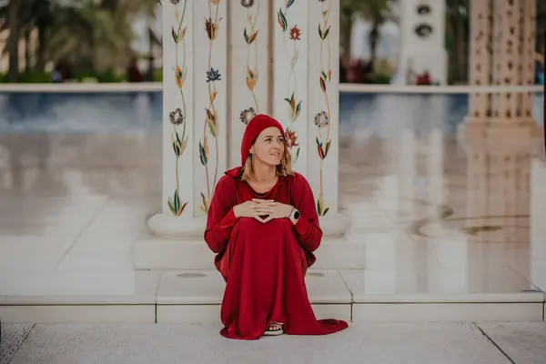 Dubai,  Unite dArab Emirates - October 19, 2019 - Seated woman in red with headscarf looking thoughtful, ornate column and reflective floor in a mosque.