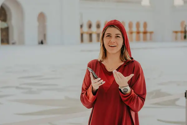 Dubai,  Unite dArab Emirates - October 19, 2019 - Woman in red with a headscarf holding a phone, smiling, and gesturing, with a mosque courtyard in the background.