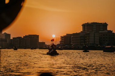 Dubai, Birleşik Arap Emirlikleri - 19 Ekim 2019 - Turuncu bir gökyüzüne karşı tekne ve bina siluetleriyle birlikte gün batımı manzarası.