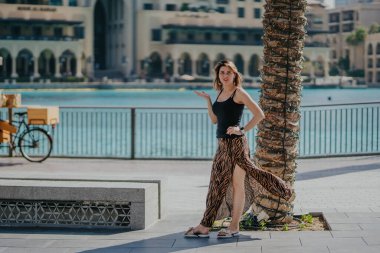 Dubai, Birleşik Arap Emirlikleri - 19 Ekim 2019 - Siyah kolsuz bluzlu ve desenli etekli bir kadın, arka planda su ve binalardan oluşan bir gövdeyle bir palmiye ağacının yanında geziniyor..