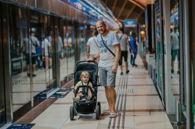 Dubai, Birleşik Arap Emirlikleri - 19 Ekim 2019 - Traşlı kafalı bir adam, içinde küçük bir çocuk olan bir bebek arabasını iterek, arka planda tren kapıları ve diğer yolcularla birlikte bir metro istasyonunda duruyor..