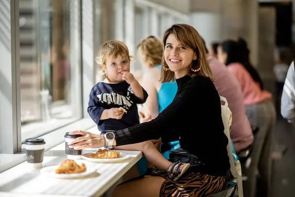 Dubai,  Unite dArab Emirates - October 19, 2019 - A woman and a child are sitting at a cafe table by a window; the woman is smiling at the camera, and the child is eating.