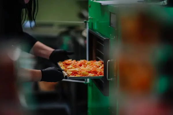 A person in black gloves is handling a tray of orange freeze-dried carrot slices in a blurred kitchen setting.