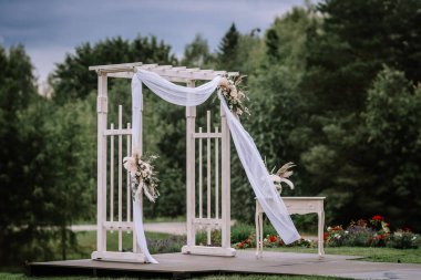 Valmiera, Letonya - 13 Ağustos 2023 - Beyaz kumaş ve çiçeklerle süslenmiş bir düğün kemeri, arka planda yeşillik olan bir bahçede ahşap bir sahneye kurulmuş..
