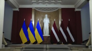 Riga Letonya - 4 Nisan 2024 - Mavi-sarı ve beyaz bayraklı bir salonda heykel, süslü merdivenler ve mimari detaylar.