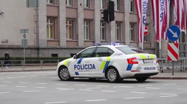 Riga Letonya - 4 Nisan 2024-Polis arabası yayalar ve bayraklarla dolu bir şehir caddesinde yanıp sönen ışıklar.