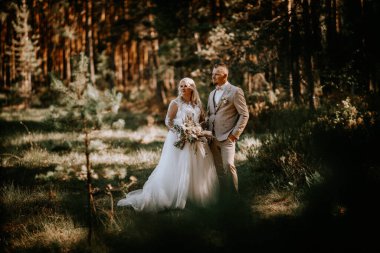 Gelin ve damat bir ormanın ortasında birlikte durur, gelin elinde buketle, ağaçların arasından süzülen yumuşak güneş ışığıyla yıkanır.