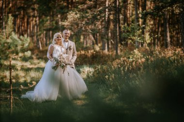Gülümseyen bir gelin ve damat, güneşli bir orman açıklığında, gelin büyük bir buket tutarken birlikte dururlar..