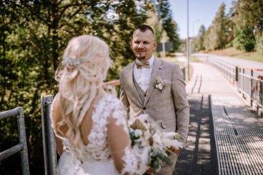 Valmiera, Letonya - 13 Ağustos 2023 - Bir damat kameradan uzak duran geline bakar, arka planda yeşillik olan güneşli bir kaldırımda.