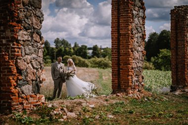 Valmiera, Letonya - 13 Ağustos 2023 - Eski tuğla sütunlar, beyaz elbiseli gelin ve arka planda tarlası olan bej takım elbiseli damat arasında bir düğün çifti duruyor..