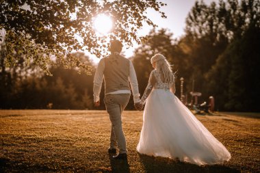 Valmiera, Letonya - 13 Ağustos 2023 - Bir gelin ve damat el ele bir tarlada yürür, arkalarından gelen ağaçların arasından güneş ışığı sahneye altın bir parıltı saçar.