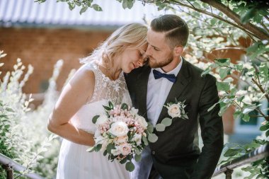 Düğün kıyafetli bir çift, yeşil ile çevrili alnına nazikçe dokunuyor, gelin bir buket tutuyor ve damat papyon takıyor..