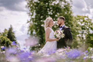 Valmiera, Letonya - 14 Temmuz 2023 - Bir düğün çifti bir çiçek tarlasında birbirlerine yakın durup parlak bir gökyüzünün altında birbirlerine bakıyorlar.