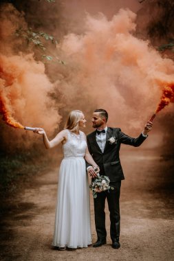 Valmiera, Letonya - 14 Temmuz 2023 - Bir düğün çifti orman yolunda, gelin solda bir duman fişeği, damat da sağda parlak turuncu dumanlar saçarak duruyor..