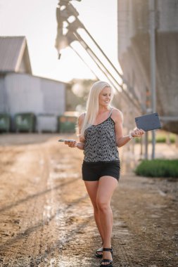 Valmiera, Latvia - August 17, 2024 - A smiling blonde woman holds a book and smartphone while standing on a farm road, with silos in the background. clipart