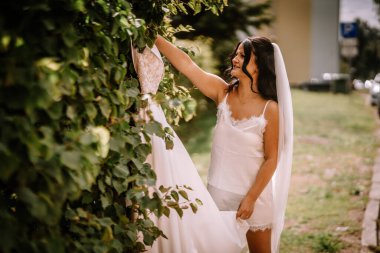 Valmiera, Letonya - 28 Temmuz 2024 - Beyaz elbiseli ve peçeli bir gelin yeşil sarmaşık arkaplanına uzanan bir gelinliğe uzanır.