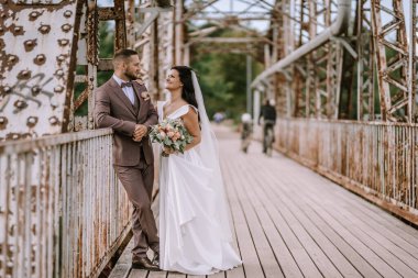 Valmiera, Letonya - 28 Temmuz 2024 - Yeni evli bir çift klasik çelik bir köprünün üzerinde duruyor, kahverengi takım elbiseli damat gelinin elini tutuyor, o da elinde buketle gülümsüyor.