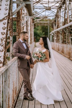 Valmiera, Letonya - 28 Temmuz 2024 - Gelin ve damat eski bir köprüde birbirlerine gülümsüyor, el ele tutuşuyorlar, gelin buketi tutuyor..
