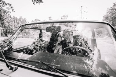 Valmiera, Latvia- 28 Temmuz 2024 - Bir gelin ve damat, ön cam tarafından ağaçların yansımalarıyla çerçevelenmiş, eski bir arabanın içinde öpüşürler.