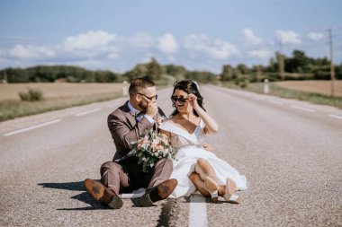 Valmiera, Letonya - 28 Temmuz 2024 - Bir gelin ve damat, neşeli ve rahat bir tavırla güneş gözlüklerini düzelterek bir yolda otururlar.