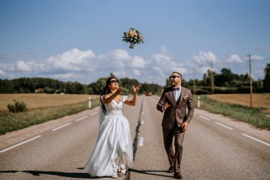 Valmiera, Letonya - 28 Temmuz 2024 - Bir gelin ve damat mavi gökyüzünün altındaki açık bir yolda heyecan içinde havaya bir buket atarlar.