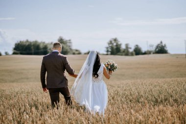 Valmiera, Letonya - 28 Temmuz 2024 Gelin ve damat bir buğday tarlasında yürüyorlar, arkadan bakıyorlar, damat bir buket çiçek tutuyor..