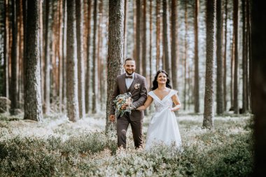 Valmiera, Letonya - 28 Temmuz 2024 Gelin ve damat el ele bir ormanda yürüyorlar, gülümsüyorlar ve ileriye bakıyorlar..