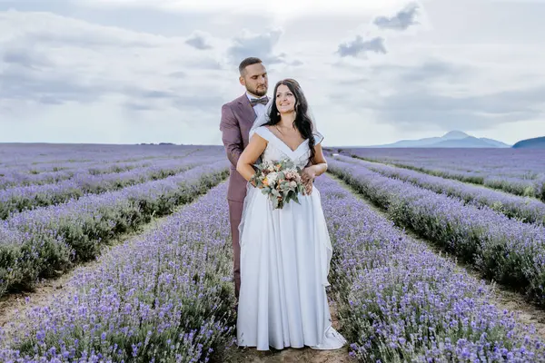 Valmiera, Latvia- July 28, 2024 - A bride and groom stand in a lush lavender field, holding hands. The groom looks at the bride lovingly while she holds a bouquet and looks away smiling.