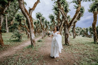 Valmiera, Letonya. 28 Temmuz 2023. Beyaz omuzsuz bir gelinlik giymiş bir gelin, eğri büğrü ağaçlarla kaplı, kirli bir patikada duruyor..