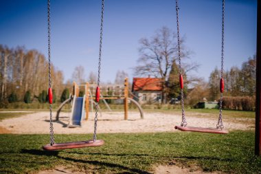Valmiera, Letonya - 21 Nisan 2024 - Arka planda açık mavi gökyüzü ve ağaçlarla dolu bir oyun parkında boş salıncaklar, barışçıl bir açık hava manzarası izlenimi uyandırıyor..